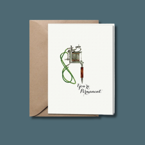 Greeting card with tattoo gun illustration and 'You're Permanent' text on a white background with a brown envelope.