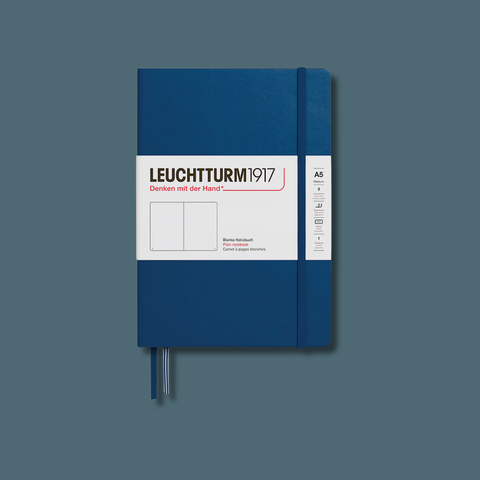 A Leuchtturm1917 A6 notebook with a dark blue cover, a white front label, and a matching blue elastic closure, shown against a solid slate blue background.