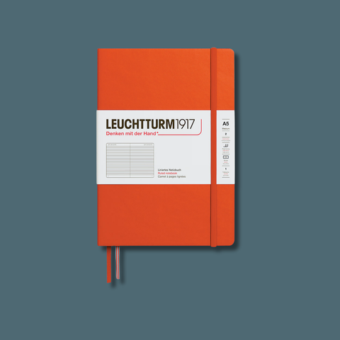 An orange hardcover notebook with a white paper band around the middle reading 'LEUCHTTURM 1917' and a vertical size/feature label on the right, shown on a muted blue-gray background.