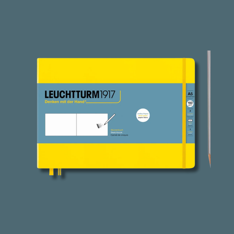 A Leuchtturm1917 A5 sketchbook with a bright yellow cover and a slate blue front label, shown on a muted blue-gray background with a pencil standing to the right.