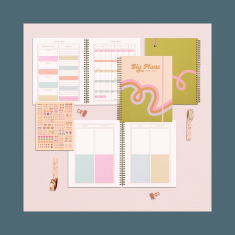 A styled flat-lay of a planner set on a pale pink background, including two open spiral-bound planner pages, a closed olive-green planner with pink swirl graphics and the words 'Big Plans', a sheet of pastel stickers, washi tape, and small clips. The outer background is a muted blue-gray.