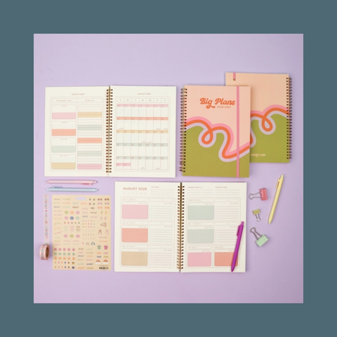 A styled flat-lay of stationery items on a lavender background, including two spiral-bound planners, two open monthly calendar pages, a sheet of sticky notes, and pens and paper clips in pastel colors.