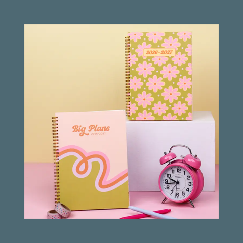 A styled desk setup featuring two spiral-bound planners, one with a pink and yellow floral cover and one with a pink and orange abstract design, alongside a pink alarm clock, a pen, and a washi tape roll on a pastel background.