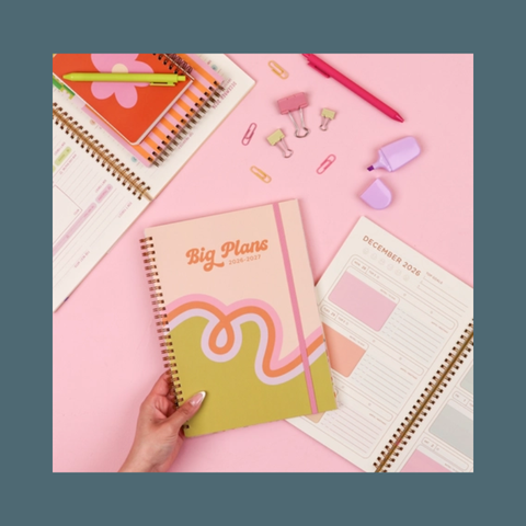 A hand holds a spiral-bound planner titled 'Big Plans 2026' with a pastel pink, orange, and yellow cover design, styled on a pink background with other stationery items.