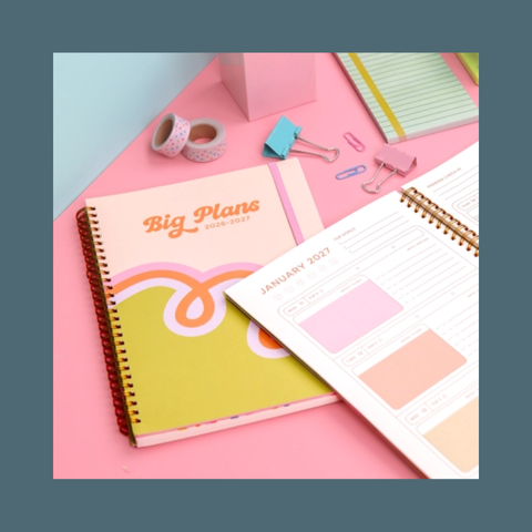 A styled flat-lay of stationery items on a pink surface, including a spiral-bound planner or notebook with the title 'Big Plans since 2021,' an open monthly calendar page, washi tape rolls, paper clips, and pastel stationery accessories.