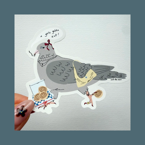 A hand holds a die-cut sticker featuring a whimsical illustration of a gray pigeon carrying a miniature woman and a gift box, set against a textured white square card with a muted blue-gray border.